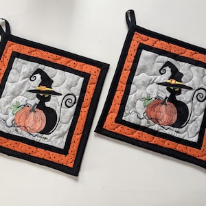 May include: Two black and orange quilted pot holders featuring a black cat wearing a witch's hat and holding a pumpkin.