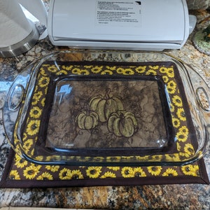 May include: A glass baking dish with handles sits on a brown and yellow quilted hot pad with a sunflower pattern. The hot pad has an embroidered design of three pumpkins.