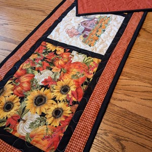 May include: A quilted table runner with a autumn theme. It features a farmhouse scene with pumpkins, sunflowers, and autumn leaves. The runner has a black and orange gingham border.