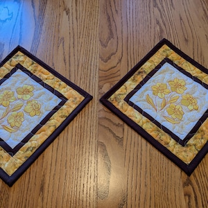 May include: Two square quilted pot holders with a floral design. The center features a light background with yellow flowers. The borders are yellow and brown, with a dark brown binding. The pot holders are placed on a wooden surface.