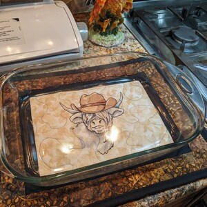 May include: A clear glass baking dish with a rectangular shape and rounded corners. Inside, a decorative insert features a Highland cow wearing a cowboy hat. The cow is depicted in a detailed, sketched style, with a tan and brown color palette.