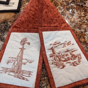May include: Two quilted pot holders with a red and white floral pattern. One pot holder features an embroidered image of a windmill with a fence and trees. The other pot holder features an embroidered image of a tractor in a field.