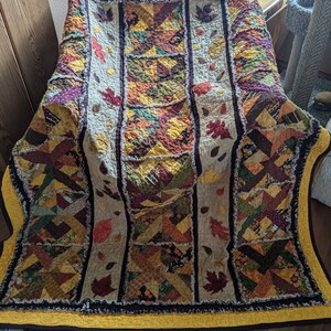 May include: A handmade quilt with an autumn theme. The quilt is made of various fabrics in shades of brown, orange, yellow, and green. It features a patchwork design with leaves and other autumn motifs. The quilt is edged with a yellow ruffle.
