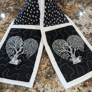 May include: A double-sided quilted oven mitt featuring a black fabric with a white star pattern at the top and black fabric with white embroidered heart-shaped trees at the bottom. The edges are finished with a light gray fabric binding.