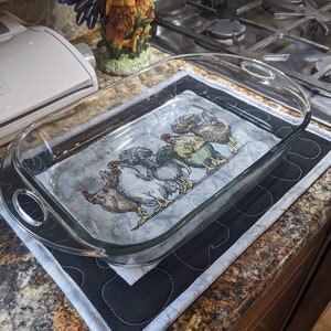 Puede incluir: Un salvamanteles acolchado en blanco y negro con fondo blanco y borde negro. El salvamanteles tiene un diseño de cinco pollos bordados. El salvamanteles está sobre una encimera con una fuente de horno de vidrio encima.