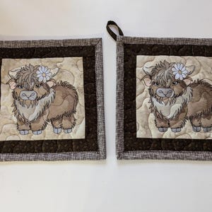 May include: Two brown and white quilted pot holders with a brown border. Each pot holder features an embroidered image of a brown and white Highland cow with a blue flower on its head.