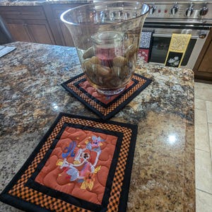 May include: Two orange and black quilted placemats with a black border. One placemat has an embroidered design of a witch's cauldron with a cat and flowers. The other placemat is empty. A glass vase with rocks and a candle inside sits on the empty placemat.