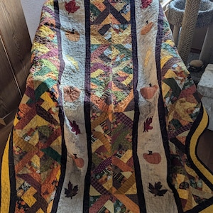 Può includere: Una trapunta patchwork con un tema autunnale. La trapunta è realizzata con vari tessuti in tonalità di marrone, arancione, giallo e verde. Presenta un motivo ripetitivo di foglie e zucche. La trapunta è bordata con un tessuto marrone scuro e un tessuto giallo.