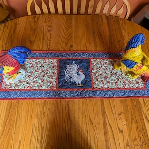 May include: A blue and red quilted table runner with a floral pattern and a white rooster design in the center. Two ceramic rooster figurines are placed on either end of the runner.