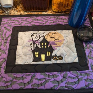 May include: A purple and black quilted Halloween-themed placemat with a black silhouette of a house with a yellow window, a full moon, and two pumpkins. The placemat is decorated with a pattern of bats.