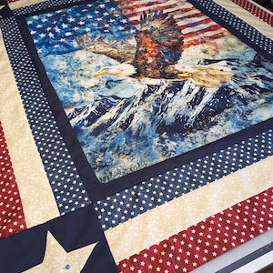May include: A patriotic quilt with an American flag design and a soaring bald eagle over a mountain range. The quilt features red, white, and blue fabrics, star patterns, and a large star accent.
