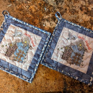 May include: Two blue and white quilted pot holders with a birdhouse design and the text "hello, friend!" embroidered on each.