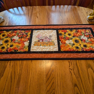 May include: A quilted table runner with a farmhouse theme. The runner features three panels with embroidered images of pumpkins, sunflowers, and a farmhouse. The runner has a black border and a red and white checkered trim.
