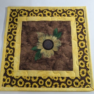 May include: A brown quilted placemat with a yellow border and a sunflower design in the center. The border is decorated with a pattern of small yellow sunflowers on a brown background.
