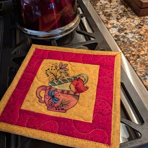 May include: A yellow and red quilted hot pad with a stitched design of a teacup with flowers and a bird. The hot pad has a red border and a yellow border.