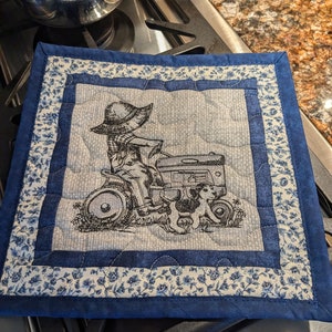 May include: Blue quilted hot pad with a white floral border. The center features an embroidered design of a person in a hat driving a tractor with two dogs following behind.