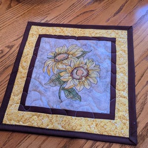 May include: A square quilted fabric mat featuring a central panel with an embroidered sunflower design. The centre is surrounded by a dark brown border and an outer yellow floral patterned border, finished with a dark brown fabric binding.