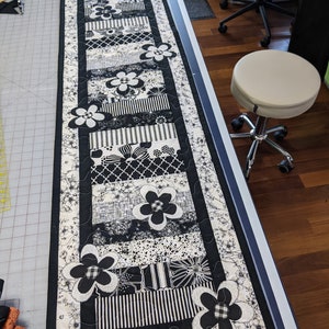 May include: Black and white table runner with a floral design. The runner is made of fabric and has a quilted texture. It features a black border and a white background with black flowers.