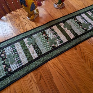 Może przedstawiać: Zielony i czarny pikowany bieżnik stołowy z wzorem koniczyny. Bieżnik wykonany jest z różnych zielonych tkanin z czarną obwódką i czarno-białym wykończeniem w kropki.