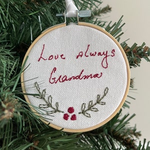 May include: A white fabric ornament with a red embroidered message that says "Love always Grandma" with green embroidered leaves and red berries.