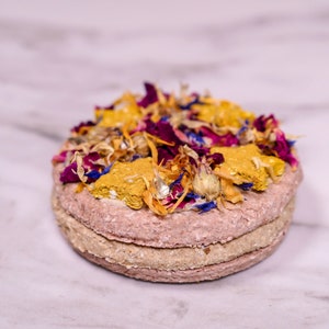 May include: A round, multi-layered dog treat topped with dried flowers and herbs. The treat is pink, beige, and yellow with a variety of dried flowers in shades of pink, purple, yellow, and blue.