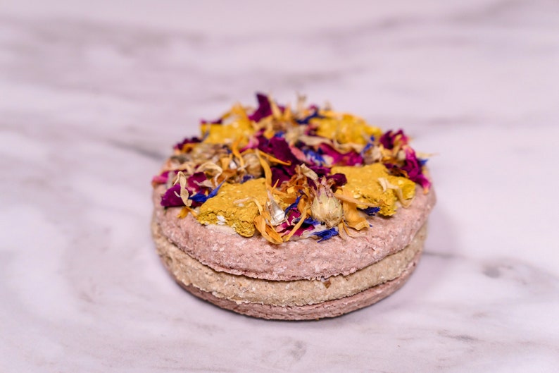 May include: A round, multi-layered dog treat topped with dried flowers and herbs. The treat is pink, beige, and yellow with a variety of dried flowers in shades of pink, purple, yellow, and blue.