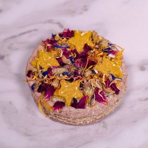 May include: A round, pink dog treat topped with dried flowers and star-shaped dog biscuits. The treat is a light pink color and has a rough texture. The flowers are a mix of pink, purple, and yellow.