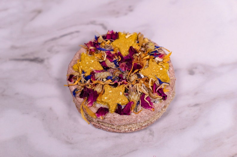 May include: A round, pink dog treat topped with dried flowers and star-shaped dog biscuits. The treat is a light pink color and has a rough texture. The flowers are a mix of pink, purple, and yellow.
