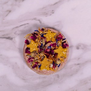May include: A round, pink, pressed flower and star-shaped treat for small animals. The treat is decorated with dried flowers in shades of pink, purple, and yellow.