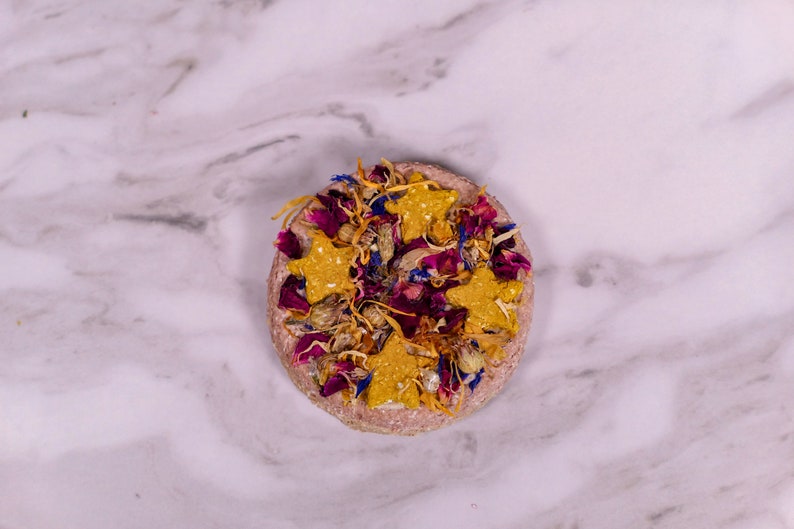 May include: A round, pink, pressed flower and star-shaped treat for small animals. The treat is decorated with dried flowers in shades of pink, purple, and yellow.