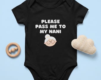 May include: A black short-sleeved baby bodysuit featuring the white text "PLEASE PASS ME TO MY NANI" above a graphic of an elderly person with glasses and a bun hairstyle, accompanied by a wooden ring toy and a small beige crocheted cloud.
