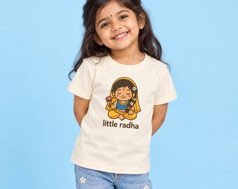 May include: A cream-colored short-sleeve t-shirt featuring a cartoon illustration of a figure in yellow and blue clothing holding a pink lotus flower, with the text "little radha" printed in brown below the graphic design on the front.