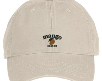 May include: A beige baseball cap featuring an embroidered design on the front. The text reads "mango season" in black, lowercase letters, with a small, orange mango fruit graphic with a dark green leaf positioned between the two words.