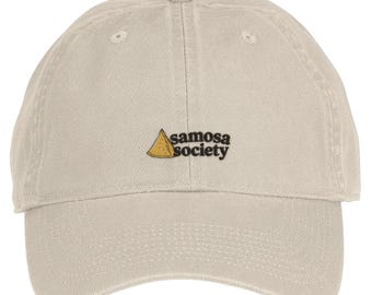 May include: A beige baseball cap featuring an embroidered graphic of a golden-brown triangular samosa next to the black text "samosa society" centered on the front panel. The cap has a curved brim and a structured crown with stitched eyelets.
