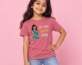 May include: A mauve short-sleeved t-shirt featuring a graphic of a person wearing a blue and gold sari with the white and yellow text "IN MY RANI ERA" printed on the front. The shirt is paired with light blue denim jeans.