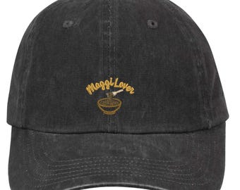 May include: A charcoal black baseball cap featuring an embroidered design on the front. The embroidery displays the text "Maggi Lover" in a yellow cursive font above a small graphic of a bowl of noodles with a fork, rendered in yellow and brown.
