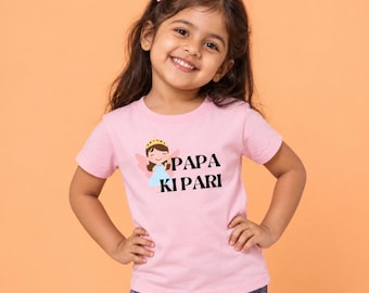 May include: A light pink short-sleeved t-shirt featuring a graphic of a cartoon fairy wearing a gold crown and a blue dress, accompanied by the black text "PAPA KI PARI" printed on the front chest area of the garment.