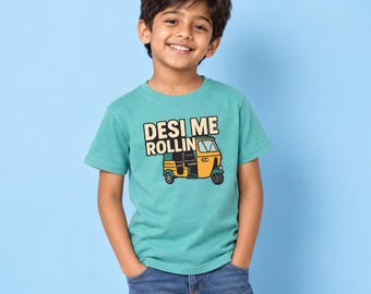 May include: A teal short-sleeve t-shirt featuring a graphic of a yellow and black auto rickshaw with the text "DESI ME ROLLIN" printed in black and cream letters above the vehicle. The shirt is paired with blue denim jeans.