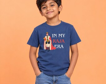 May include: A navy blue short-sleeved t-shirt featuring a graphic of a king wearing a crown and a red cape, accompanied by the white and red text that reads IN MY RAJA ERA. The shirt is paired with blue denim jeans, creating a casual look.