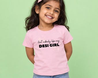 May include: A light pink short-sleeved t-shirt featuring the black text "Aint nobody like my DESI GIRL" printed on the front, accented with small star graphics. The shirt is paired with blue denim jeans and a small pink hair clip.