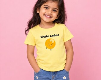 May include: A bright yellow short-sleeved t-shirt featuring a graphic of a round orange sweet with a smiling face and a cashew on top, accompanied by the black text Little Ladoo, paired with blue denim jeans decorated with white flower patterns.