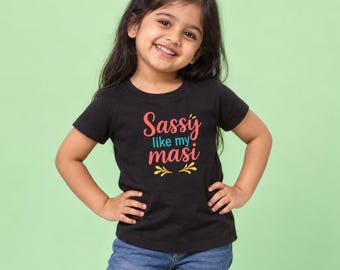 May include: A black short-sleeved t-shirt featuring the text Sassy like my masi printed in coral and teal, accented with yellow decorative flourishes. The shirt is paired with blue denim jeans, creating a casual outfit for a young child.