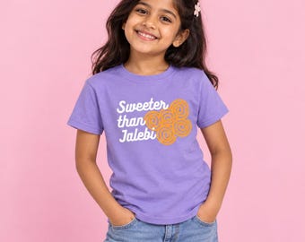May include: A light purple short-sleeve t-shirt featuring the white text Sweeter than Jalebi and an orange graphic of five spiral-shaped jalebi sweets. The shirt has a crew neckline and is paired with blue denim jeans.
