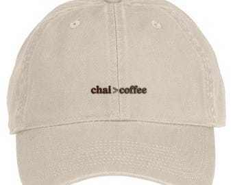 May include: A beige baseball cap featuring the embroidered text "chai > coffee" in dark brown thread. The cap has a six-panel construction with a curved brim, a button at the top, and stitched eyelets for ventilation on each panel.