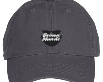 May include: A charcoal gray baseball cap featuring an embroidered black graphic of a beard and sunglasses with the white text Brown Munde centered on the front. The cap has a curved brim, a button on top, and stitched eyelets for ventilation.
