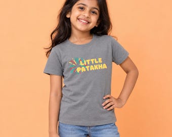 May include: A grey short-sleeved t-shirt featuring the text LITTLE PATAKHA in yellow letters with a colorful firework graphic to the left. The shirt has a crew neckline and is made of a cotton-blend fabric, paired with blue denim jeans.