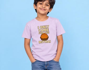 May include: A light purple short-sleeved t-shirt featuring a graphic of an orange dessert in a brown wrapper. The text above and below the dessert reads I ONLY CAME FOR THE MITHAI in yellow block letters with a dark outline on the front of the shirt.
