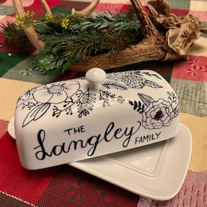 Puede incluir: Mantequera de cerámica blanca con tapa, decorada con diseños florales negros. La tapa presenta las palabras "The Langley Family" en una elegante escritura. La mantequera se asienta sobre una base rectangular blanca a juego.