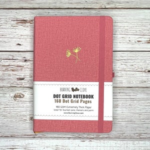 May include: A pink dot grid notebook with a gold floral design on the front cover. The notebook has a white label with the text "BURNING CLOVE DOT GRID NOTEBOOK 160 Dot Grid Pages 160 GSM Extremely Thick Paper Great for fountain pens, markers and paint www.BurningClove.com".