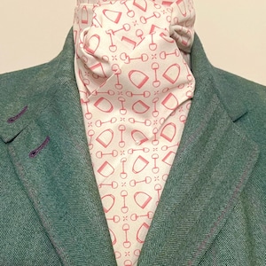May include: A teal green herringbone blazer with purple stitching and a white ascot. The ascot features a repeating pattern of pink equestrian bits and stirrups. The blazer is buttoned at the top.